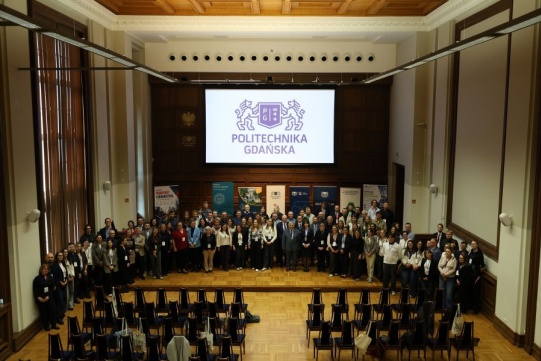 Zdjęcie grupowe uczestników konferencji „Transport i Urbanistyka 2026” wykonane w auli Politechniki Gdańskiej. Kilkadziesiąt osób stoi na scenie na tle dużego ekranu z logotypem uczelni oraz roll-upów organizatorów i partnerów wydarzenia.